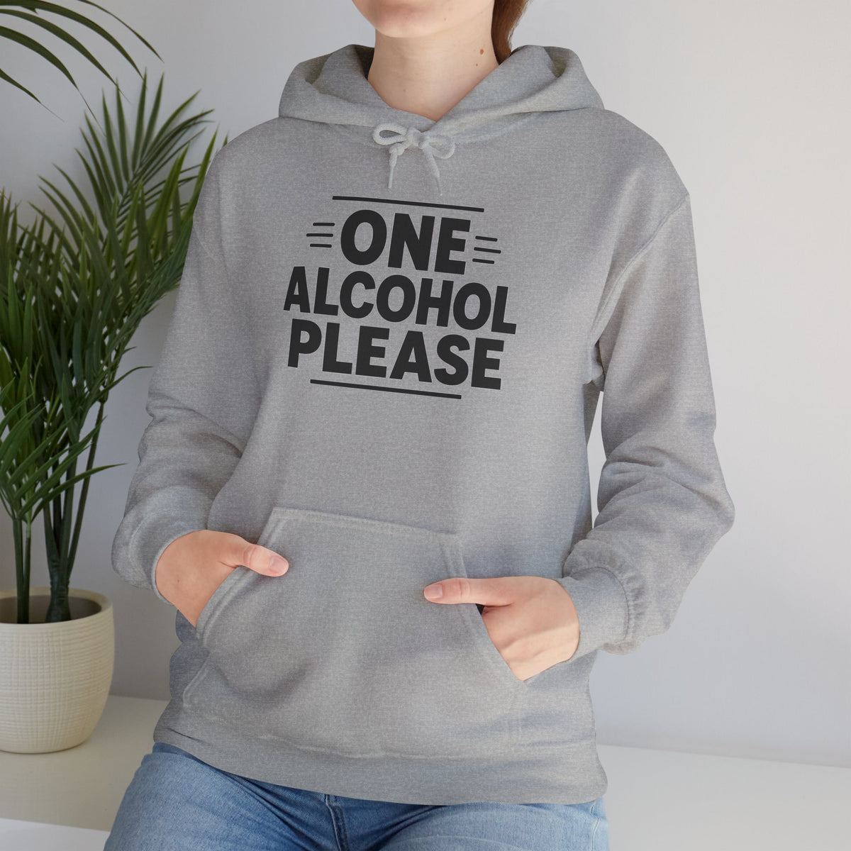 One Alcohol Please funny drinking hoodie, Sport Grey, cropped torso close-up with hands in the front pouch pocket and a houseplant behind