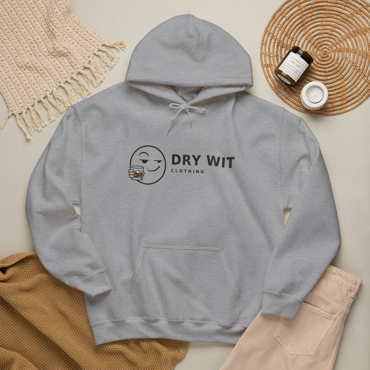 Dry Wit Clothing logo hoodie with winking emoji holding a whiskey glass, Sport Grey, flat lay styled with a knit throw, candle and tan trousers
