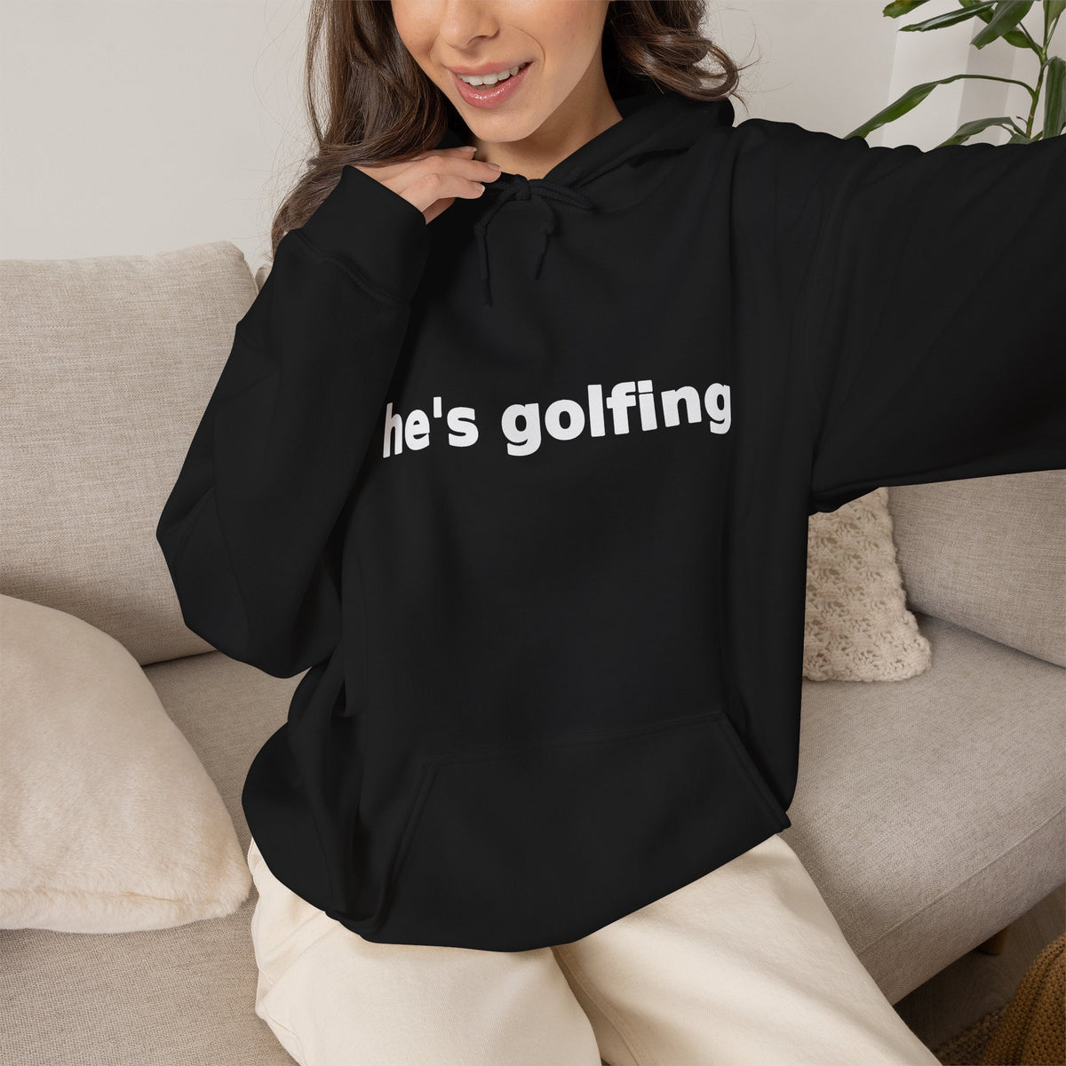Golf Widow he's golfing funny golf hoodie, Black, worn by a model seated on a sofa