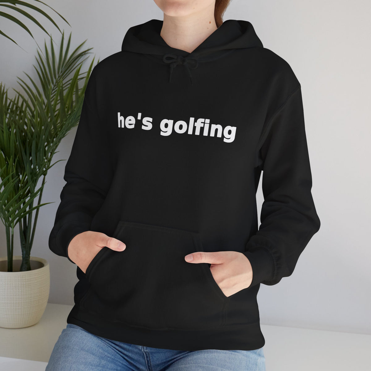 Golf Widow he's golfing funny golf hoodie, Black, cropped torso close-up with hands in the front pouch pocket and a houseplant behind