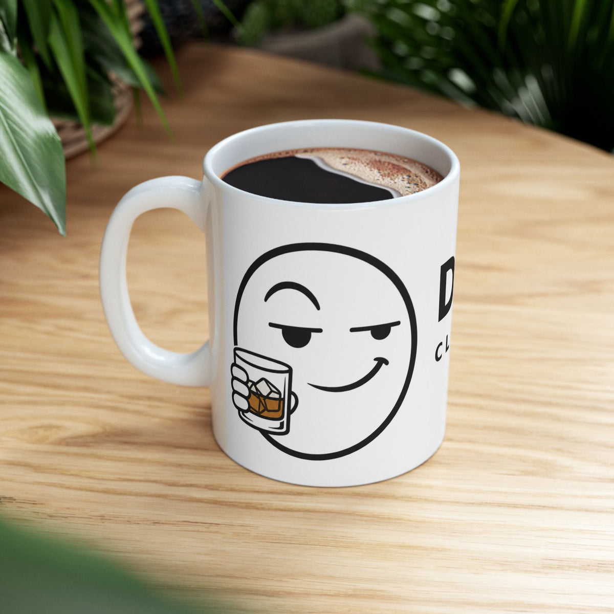 Dry Wit Logo Mug, white ceramic, lifestyle shot on a wooden table beside houseplants, filled with black coffee, handle on the left