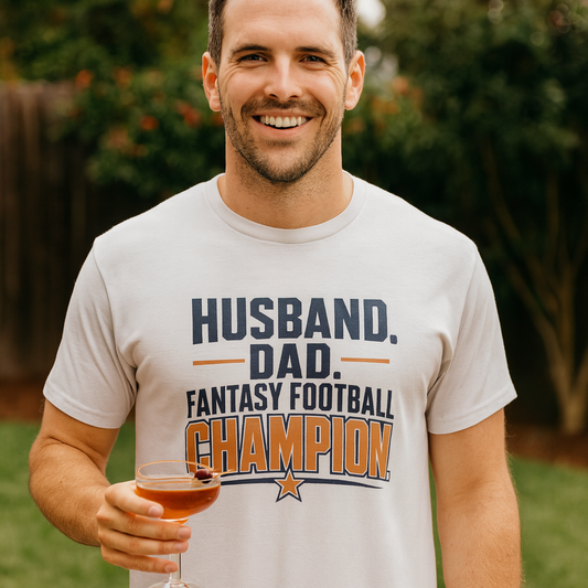 Smiling man in a white Husband Dad Fantasy Football Champion shirt holding a cocktail in a backyard