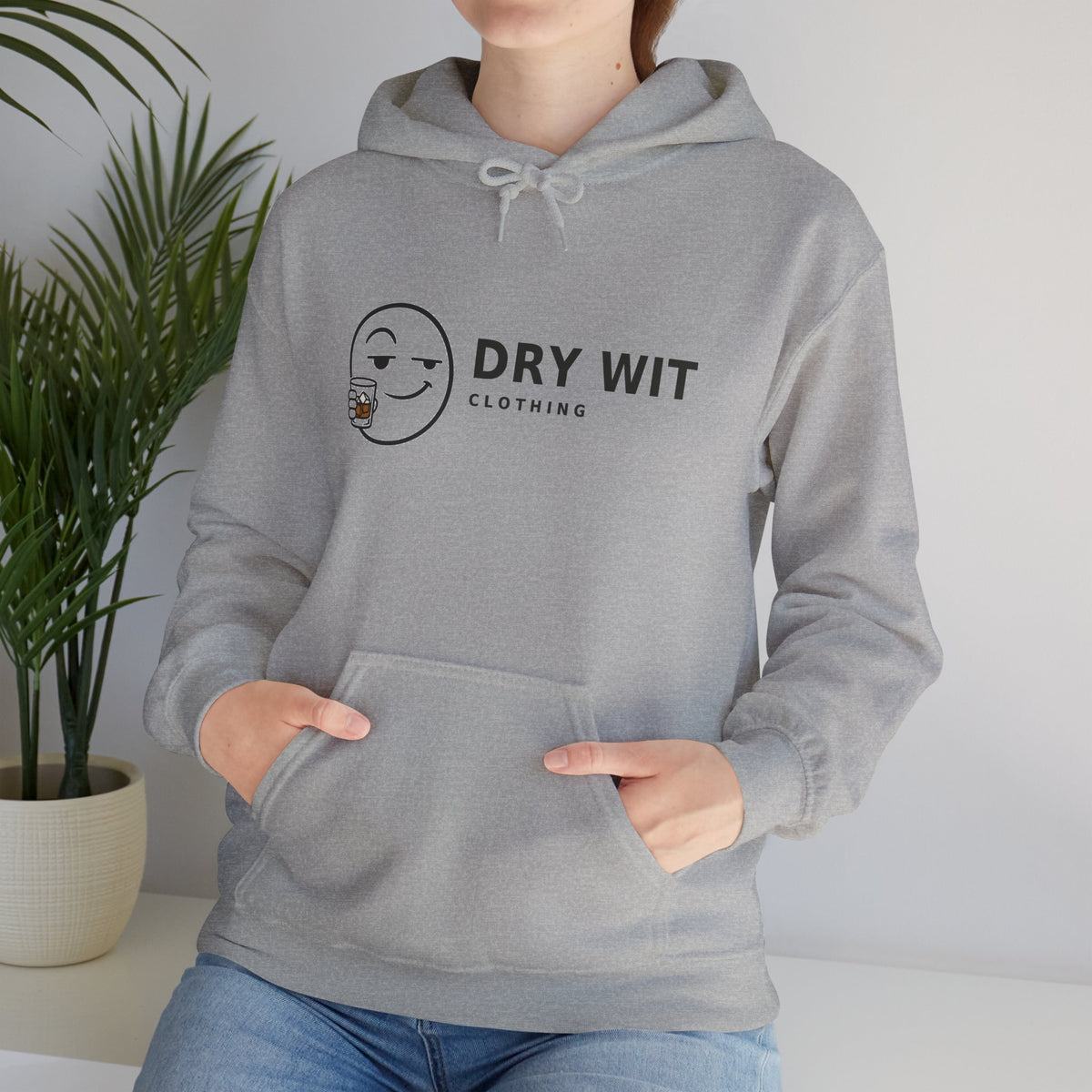 Dry Wit Clothing logo hoodie with winking emoji holding a whiskey glass, Sport Grey, cropped torso close-up with hands in the front pouch pocket and a houseplant behind