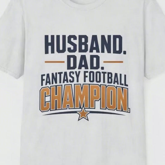 Video of a man standing proudly holding up a small trophy with a big grin, wearing a white t-shirt with navy and amber text reading Husband Dad Fantasy Football Champion