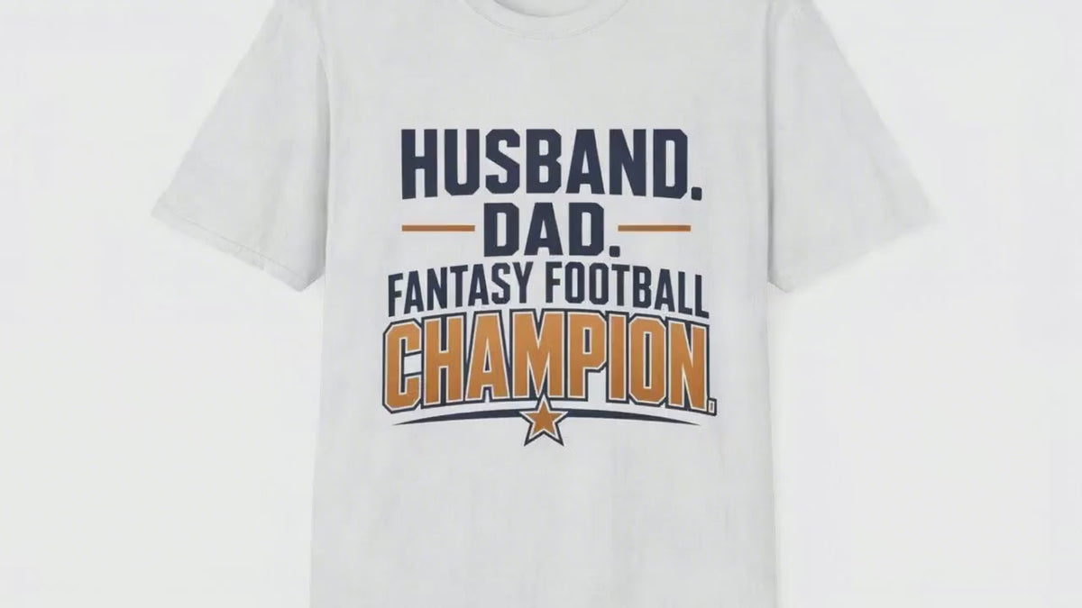 Video of a man standing proudly holding up a small trophy with a big grin, wearing a white t-shirt with navy and amber text reading Husband Dad Fantasy Football Champion